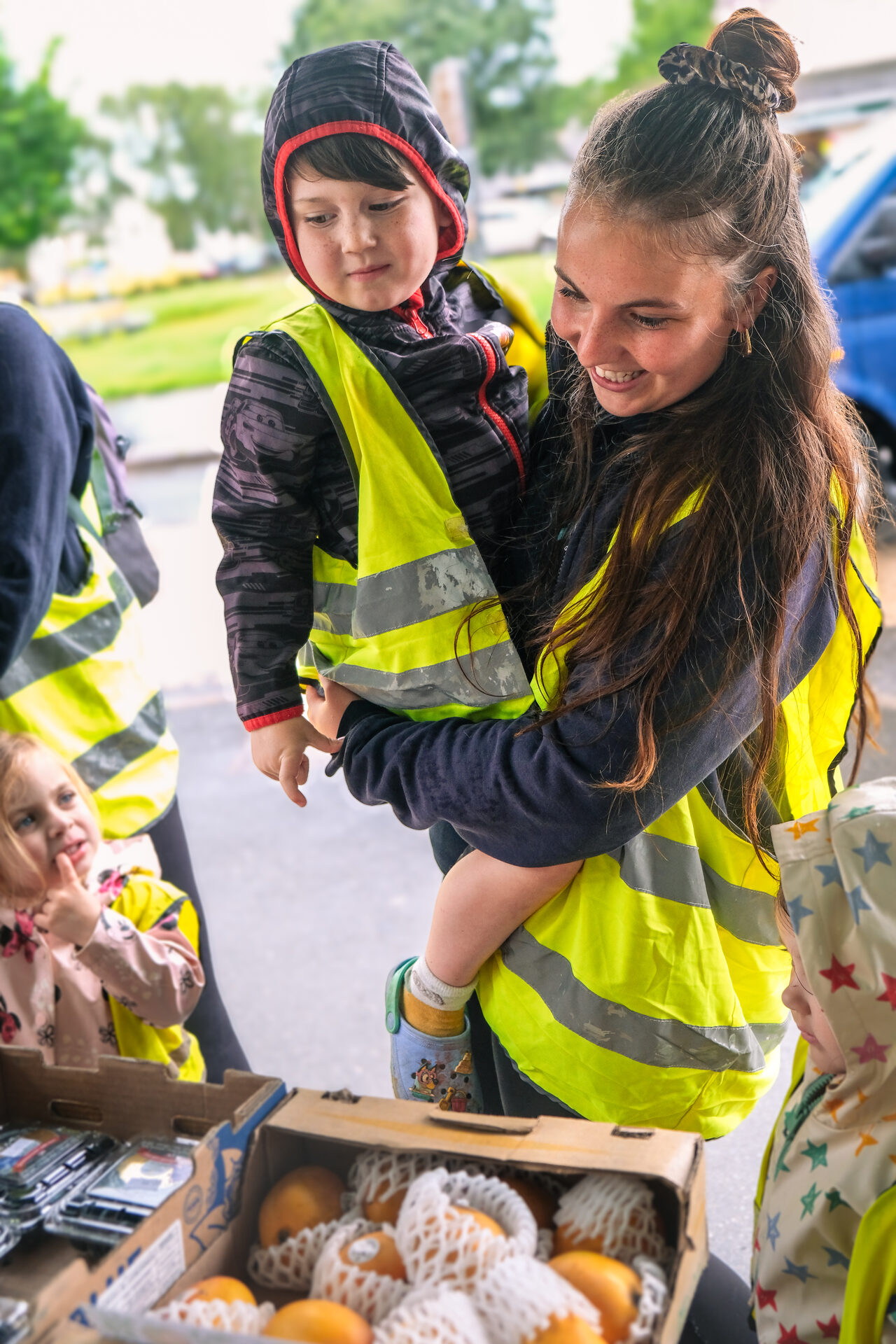 A nursery team member carrying a child in a high-visibility vest while exploring a market stall